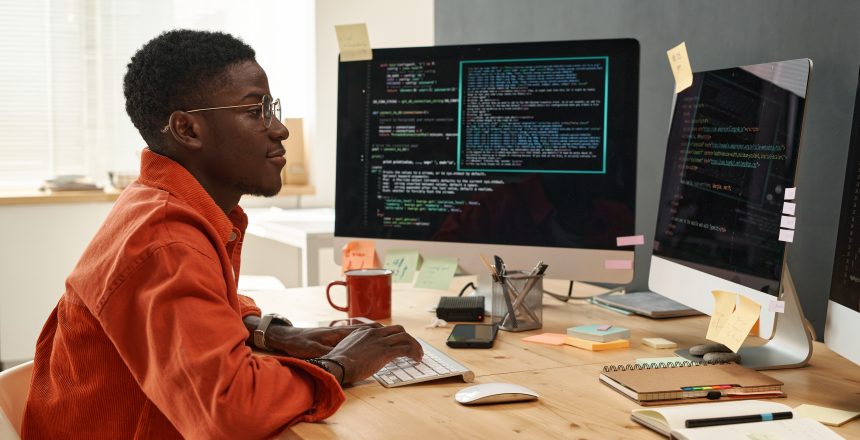 Young serious IT engineer in smart orange shirt looking at coded data on computer screens while typing on keyboard by workplace